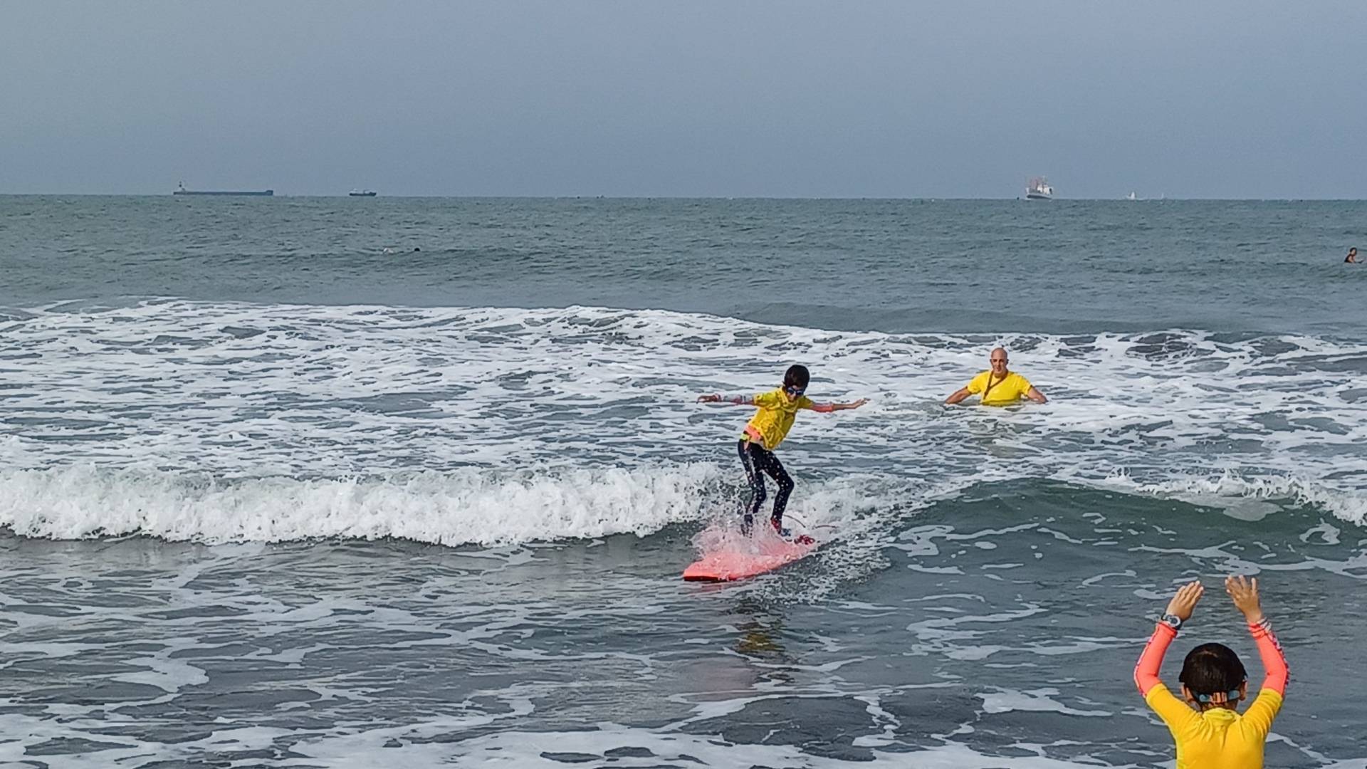 Child stands up on a surfboard. Coach in the background. Child supporter celebrating in the foreground.