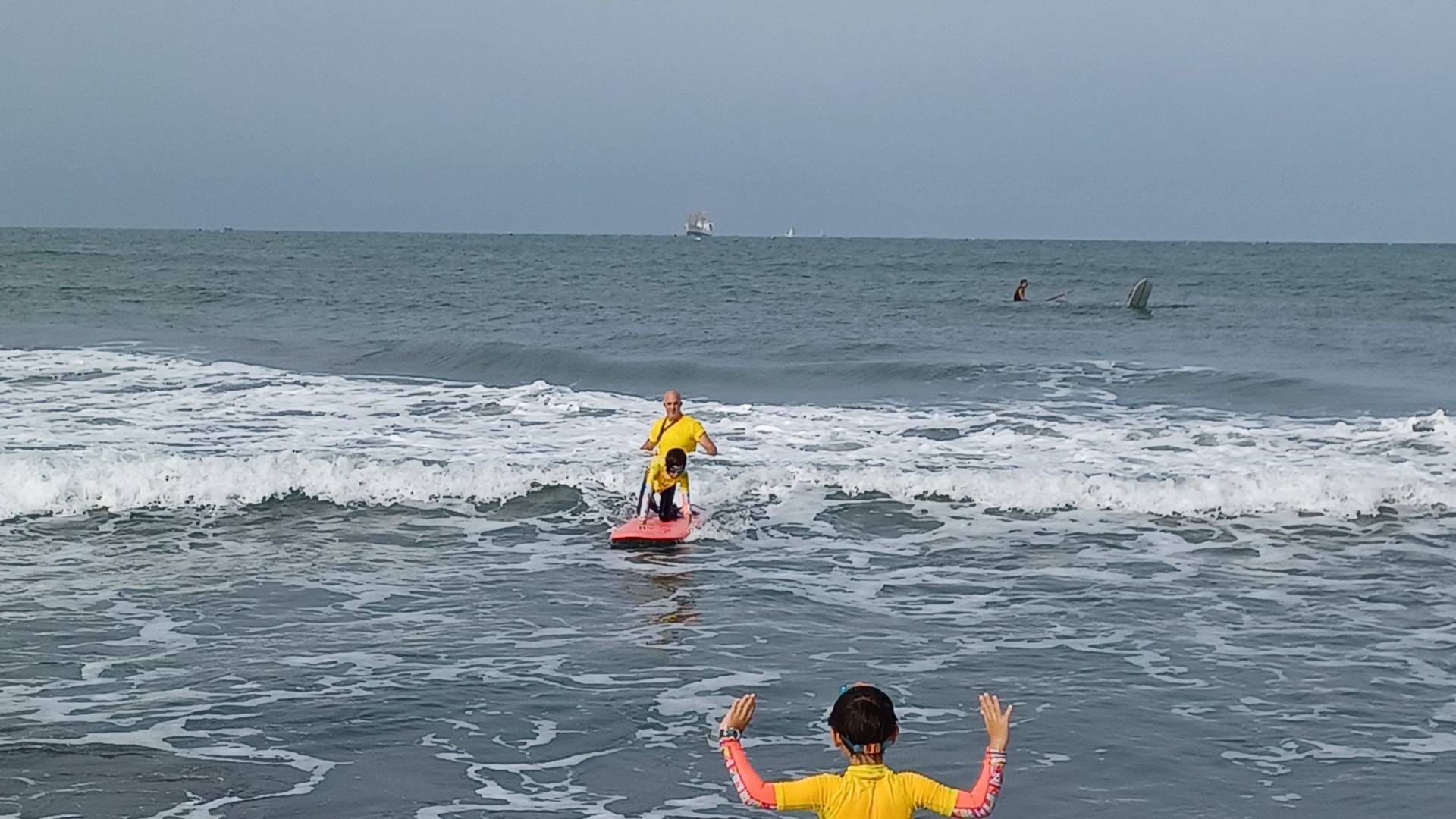 Child attempting to stand up on a surfboard. Coach in the background. Child supporter in the foreground.